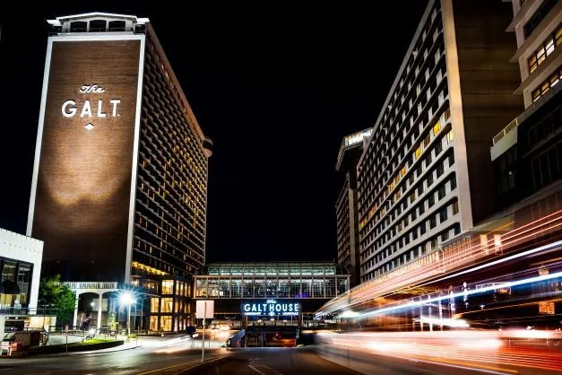 Night view of the Galt House Hotel buildings with long exposure light trails from passing traffic, emphasizing the illuminated signage and urban setting.