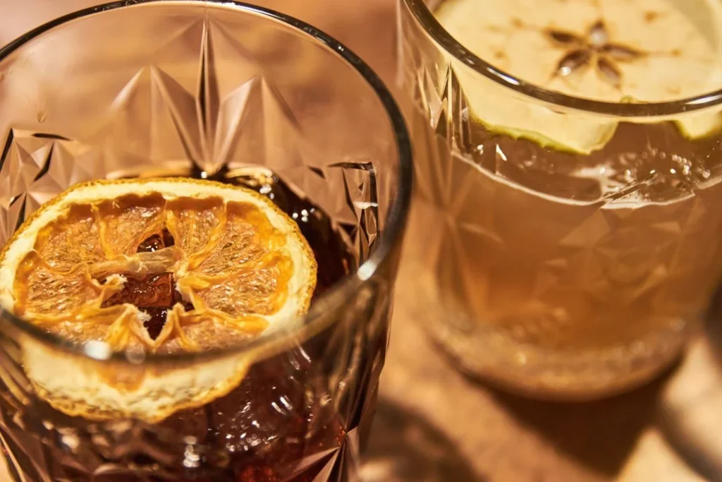 Two crystal glasses with cocktails, each topped with a dried citrus slice, on a wooden surface. The image emphasizes the drinks' garnishes and glass detail.