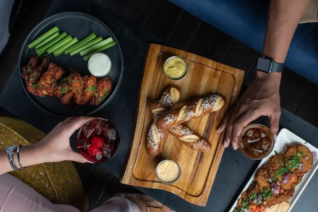 Overhead view of two people sharing appetizers, including chicken wings, pretzel sticks, and breaded cutlets, with celery, dips, and cocktails. The filename suggests a restaurant or food-related context, possibly Kriech-Higdon establishment.