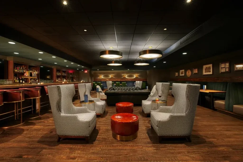 Modern hotel lounge with houndstooth chairs, round red ottomans, a tufted black sofa, and a bar area. Elegant lighting and decor suggest an upscale atmosphere, likely the Galt House Jockey Silks interior.